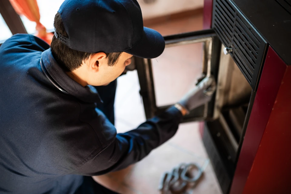 Photographie réaliste d'un technicien certifié en train d'intervenir sur un poêle à granulés ou un poêle à bois en intérieur résidentiel. Le technicien porte des vêtements professionnels, utilise des outils d'entretien ou de dépannage, et inspecte l'appareil. L'ambiance hivernale ou la présence d'un client aux alentours peuvent renforcer le contexte d'urgence et de service rapide. La photo doit montrer une intervention proactive et professionnelle, transmettant la confiance et la compétence.